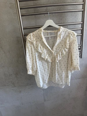 Vit prickig blus med volang - Supersöt vit blus med svarta prickar och volangdetaljer längs ringningen. Blusen har trekvartsärm och knappar framtill. Perfekt för dig som gillar romantisk och trendig stil.