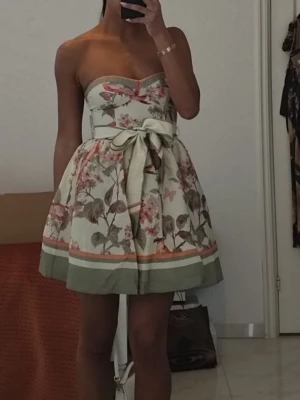 Blommig axelbandslös kortklänning från h&m - Supersöt axelbandslös kortklänning med blommigt mönster i grönt, rosa och beige. Klänningen har en markerad midja med stor rosett framtill och en vid kjol med grön kant nedtill. Perfekt för sommarens alla tillfällen!