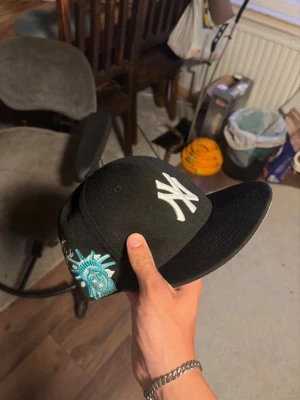 Svart New Era keps med NY-logga - Säljer en svart New Era keps med klassisk New York Yankees-logga i vitt framtill och broderad Frihetsgudinnan på sidan. Kepsen har platt skärm och sticker ut med coola detaljer. Perfekt för dig som vill ha en streetstyle-look. Storlek är 57.7 cm  eller 7 1/4