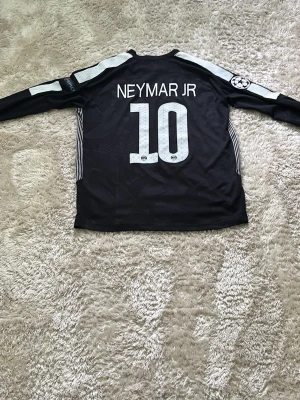 PSG Neymar Jr långärmad matchtröja - Svart långärmad Paris Saint-Germain matchtröja från Nike med Neymar Jr och nummer 10 på ryggen. Tröjan har vita detaljer, Fly Emirates-logga på bröstet och Champions League-märke på ärmen. Perfekt för fotbollsfans!