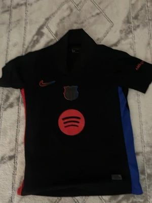 FC Barcelona svart matchtröja Nike - Svart FC Barcelona matchtröja från Nike med röd och blå detalj på sidorna. Tryck med Spotify-logga framtill och UNHCR-logga bak. Kortärmad med klubbmärke och Nike-logga på bröstet. Perfekt för fotbollsträning eller som supportertröja.