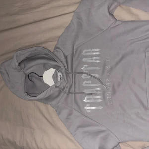 Trapstar dress - Säljer en grå hoodie från Trapstar med stort vitt tryck på bröstet och snörning i huvan. Tröjan har en klassisk passform och är tillverkad i mjukt material, perfekt för chill dagar. Snygg streetwear-vibe och passar till allt.