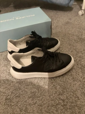 Svarta Philippe Model sneakers - Snygga svarta sneakers från Philippe Model med vita detaljer och vit sula. Skorna har en klassisk låg design med svarta skosnören och diskret logga på sidan. Tillverkade i läder för en stilren och modern look.