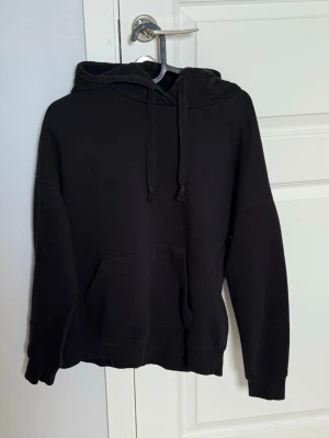Svart hoodie med ficka och huva - En klassisk svart hoodie med stor magficka och justerbar huva med snören. Hoodien har ribbade muddar vid ärmslut och nederkant. Perfekt för en avslappnad och stilren look. Materialet känns mjukt och bekvämt, troligen bomullsblandning.