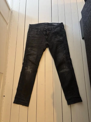 Replay anbass jeans med slitningar - Säljer ett par svarta jeans från Replay med snygga slitningar och tvättad look. Modellen är slim storlek 32