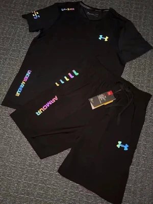 Svart träningsset från Under Armour - Säljer ett par svart träningsset från Under Armour med färgglada loggor och textdetaljer. Shortsen har snörning i midjan och är tillverkade i ett lätt och sportigt material som passar perfekt för träning eller chill. Snygg och modern design med regnbågsfärgade detaljer. Storlek S, helt ny 