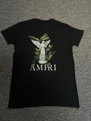 Svart AMIRI t-shirt med tryck - Svart t-shirt från AMIRI med coolt tryck av en ängel och gröna blad på ryggen samt AMIRI-logga. Framsidan har AMIRI-tryck med färgstänk. Klassisk passform och korta ärmar, perfekt till jeans eller shorts. Tillverkad i mjuk bomull för skön känsla.