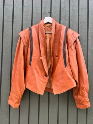 Vintage brun skinnjacka med markerade axlar  - Unik brun skinnjacka i retrostil med markerade axlar och svarta detaljer längs axlarna. Kortare modell med bred krage och två knappar framtill. Snyggt skuren för en cool siluett och perfekt för dig som vill sticka ut.