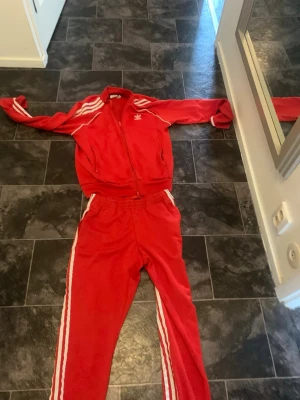 Röd Adidas träningsdress - Säljer en klassisk röd Adidas träningsdress med vita ränder längs ärmar och ben. Jackan har dragkedja och två fickor, byxorna har resår i midjan och muddar vid benslut. Ikonisk Adidas-logga på bröstet. Perfekt för sport eller chill.