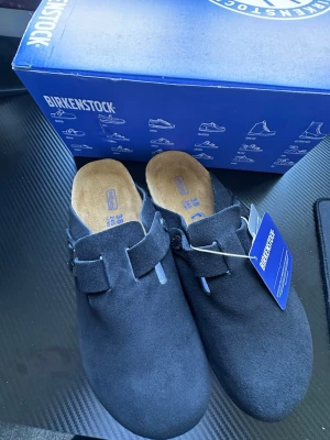 Svarta Birkenstock Boston tofflor - Helt nya Stilrena svarta Birkenstock Boston tofflor i mocka med justerbart spänne och klassisk korksula. Perfekta för dig som gillar en clean och bekväm look. Innersulan är mjuk och följer fotens form för extra komfort.