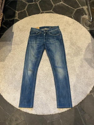 Blå slim fit jeans dondup - Säljer ett par blå slim fit jeans med snygga slitningar och tvättade detaljer. Klassisk femficksmodell med dragkedja och knapp i midjan. Jeansen har en smal passform och är tillverkade i denim. Perfekta för en avslappnad och trendig look.