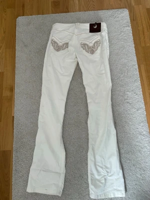 Vita jeans med broderade fickor - Säljer ett par vita jeans med snygga broderade detaljer på bakfickorna. Jeansen har låg midja, utsvängda ben och dekorativa sömmar framtill. Perfekta för dig som vill sticka ut lite extra med coola detaljer.