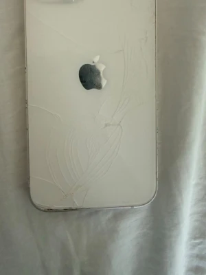Vit iPhone med glasbaksida - Säljer en vit iPhone13 med  glasbaksida med sprickor och klassisk Apple-logga. Telefonen har rundade hörn och metallram. Perfekt för dig som gillar enkel och snygg design. Har även några sprickor på framsidan