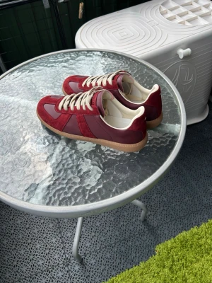 Bordeaux/burgundy gats från Maison Margiela - Säljer ett par bordeauxfärgade gats från Maison Margiela. Skorna har en kombination av mocka och läder, vita snören och beige yttersula. Klassisk låg modell med retrovibbar och snygga detaljer. Perfekta för dig som gillar stilrena men unika sneakers.
