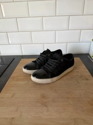 Lanvins uk7 stl 41 - Snygga svarta sneakers med glansiga lackdetaljer på tån och klassisk vit sula. Skorna har svarta skosnören och metallhål för snörningen. Ovandelen är i mockaimitation och tån i blankt material för en cool kontrast. Storlek 41-42 uk7. Väldigt bra skick bara smutsiga på bilden. box finns också hör av er vid funderingar