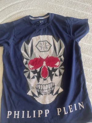 Blå Philipp Plein t-shirt med döskalle - Cool blå t-shirt från Philipp Plein med stort döskalletryck framtill i vitt, svart och rött. T-shirten har rund halsringning och korta ärmar. Märkesnamnet är tryckt längs nederkanten. Perfekt för dig som gillar edgy streetstyle.