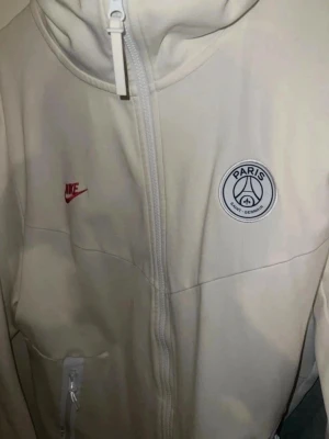 Vit PSG hoodie från Nike - Snygg vit hoodie från Nike med PSG-logga på bröstet och röd Nike Swoosh. Hoodien har hel dragkedja, fickor med dragkedja och en modern passform. Perfekt för dig som gillar fotboll och streetstyle.
