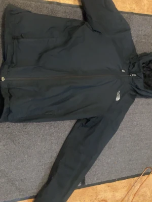 Svart vindjacka från The North Face - Svart vindjacka från The North Face med huva och dragkedja framtill. Jackan har två sidofickor med dragkedja och en diskret logga på bröstet. Perfekt för blåsiga dagar och enkel att matcha med olika outfits.
