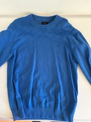 Blå stickad tröja från GANT - Snygg blå stickad tröja från GANT med rund halsringning och diskret broderad logga på bröstet. Tröjan har ribbade muddar vid ärmslut och nederkant samt en klassisk passform. Perfekt för dig som gillar stilrena och tidlösa plagg.