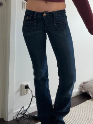 Mörkblå bootcut jeans med fickor - Snygga mörkblå bootcut jeans med låg midja och klassiska fem fickor. Jeansen har en knapp- och dragkedjegylf samt dekorativa sömmar. Perfekta för en trendig och avslappnad look. Storlek W25 L34