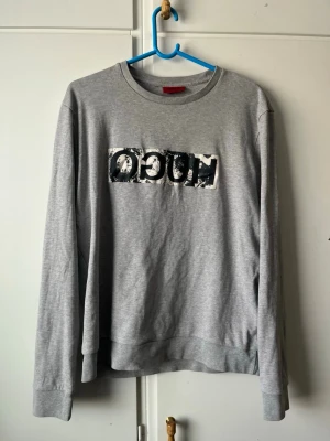 Grå sweatshirt från Hugo Boss - Snygg grå sweatshirt från Hugo Boss med tryckt logga i svart och vitt på bröstet. Tröjan har rund halsringning, långa ärmar och är gjord i mjukt bomullsmaterial. Perfekt för en avslappnad och trendig look.