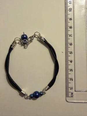 Svart armband med blå pärlor - Stilrent armband med svart band och blå pärlor i olika storlekar. Armbandet har silverfärgade metalldetaljer och ett justerbart lås. Perfekt accessoar för att ge din outfit en cool touch.