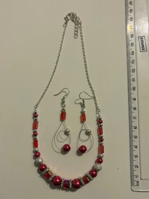 Rött pärlhalsband med matchande örhängen - Snyggt halsband med röda och silvriga pärlor samt röda glaspärlor på en tunn silverfärgad kedja. Matchande örhängen med liknande pärlor och detaljer. Perfekt för att ge din outfit lite extra färg och stil. Nickelfria