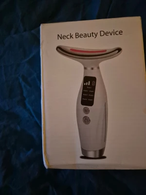 Elektrisk Neck Beauty Device - Säljer en elektrisk Neck Beauty Device för hudvård av halsen. Den är vit med silverdetaljer och har en T-formad design med digital display och flera knappar för olika lägen. Perfekt för att massera och vårda huden på halsen.