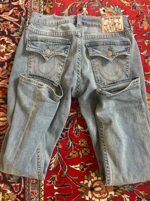 Ljusblå True Religion jeans - Säljer ett par ljusblå jeans från True Religion med raka ben och klassiska bakfickor med lock och knapp. Jeansen har en avslappnad passform och är tillverkade i denim med snygga detaljer på fickorna. Perfekta för en cool och avslappnad stil.