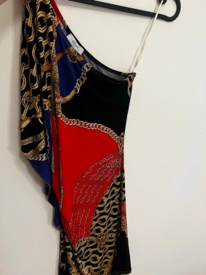 Oneshoulder klänning med kedjemönster - Snygg oneshoulder klänning från Gina Tricot med färgstarkt kedjemönster i svart, rött, blått och guld. Klänningen har en asymmetrisk axel och är tillverkad i ett mjukt, stretchigt syntetmaterial. Perfekt för dig som vill sticka ut med en unik look.