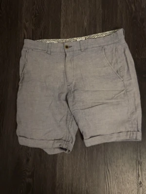 Ljusgrå shorts från Jack & Jones - Snygga ljusgrå shorts från Jack & Jones med regular fit. Shortsen har klassisk design med två bakfickor med knappar och uppvikta benslut. Perfekta för varma dagar och enkla att matcha med olika outfits.