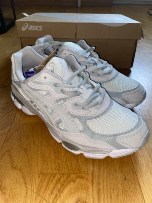 Asics nyc gel - Snygga vita och ljusgrå sneakers från Asics med klassisk mesh- och syntetöverdel. Skorna har en sportig siluett, tydliga Asics-loggor på sidorna och en chunky sula för extra komfort. Perfekta för dig som gillar stilrena och fräscha sneakers.