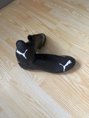 Svarta Puma fotbollsskor med högt skaft - Säljer ett par svarta fotbollsskor från Puma med högt skaft och vita loggor. Skorna har en strömlinjeformad design och ovandel i syntetmaterial för bra bollkontroll. Perfekta för dig som vill ha stilrena och funktionella skor på planen. skorna är oanvända helt nya utan prislapp 