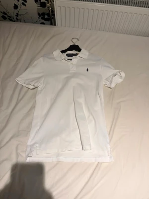 Vit pikétröja från Polo Ralph Lauren - Klassisk vit pikétröja från Polo Ralph Lauren med broderad marinblå logga på bröstet. Tröjan har korta ärmar, krage och knappar framtill. Perfekt för en clean och stilren look.