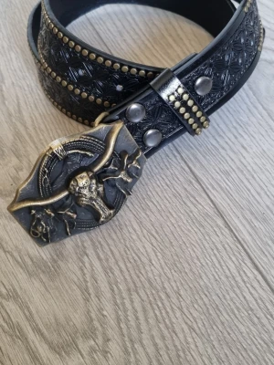 Svart Cowboy Bälte med western-spänne 110 CM - Snyggt svart bälte med präglat mönster och nitar längs kanterna. Spännet är i simulerat antikguld och har en cool western-design med tjurhuvud och repdetaljer. Perfekt för dig som vill sticka ut med en unik accessoar.