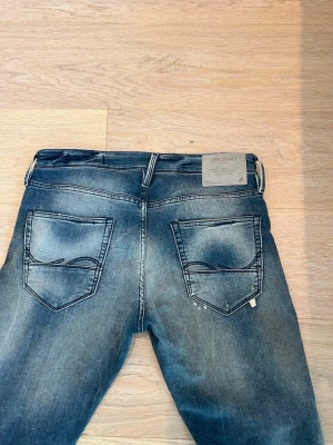Blå slitna jeans från Jack & Jones - Säljer ett par blå jeans från Jack & Jones Originals med slitningar, patchdetalj och cool tvätt. Jeansen har raka ben, fem fickor och klassisk knappgylf. Materialet är bomull med en skön känsla och passformen är smal. Perfekta för en avslappnad streetstil.