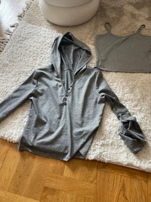 Grå långärmad tröja med knappar och huva - Säljer en snygg grå tröja med lång ärm och djup v-ringning med knappar framtill. Tröjan har en stor huva och är gjord i ett mjukt, stretchigt material som känns skönt mot huden. 
