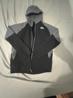 Svart och grå vindjacka från The North Face - Säljer en snygg vindjacka från The North Face i svart och grått. Jackan har huva, dragkedja framtill och logotyp på bröstet och ryggen. Perfekt för blåsiga dagar och outdoor-aktiviteter. Materialet är lätt och funktionellt.