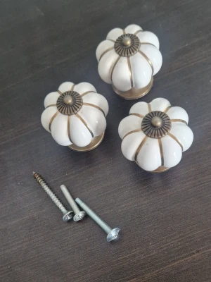 Knoppar i keramik - Tre vita knoppar i keramik med blomformad design och detaljer i mässingsfärgad metall. Perfekta som handtag till lådor eller skåp. Skruvar för montering medföljer (tyvärr ej original skruvarna). Ger en vintage och stilren känsla till inredningen.