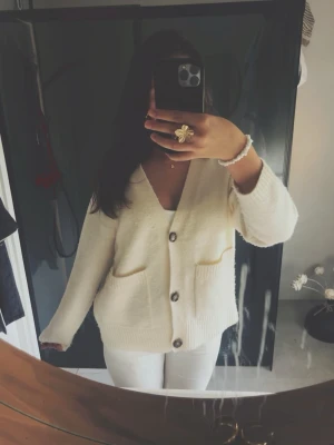 Krämvit kofta med stora knappar - Mysig krämvit kofta med v-ringning och stora knappar framtill. Koftan har två fickor och är stickad i ett mjukt material. Perfekt för lager-på-lager och chill dagar. Använd och lite nopprig, men går säkert att ta bort med maskin.