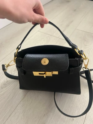 Svart handväska med gulddetaljer - Stilren svart handväska i slätt och strukturerat läder med guldfärgade metalldetaljer. Väskan har både ett kort handtag och en lång justerbar axelrem som kan tas av. Lock med vridlås framtill och elegant, kompakt form.