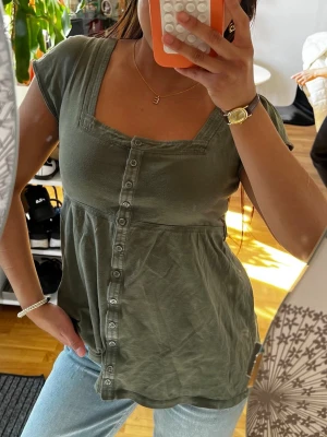 Grön topp med knappar framtill - Snygg grön topp med korta ärmar och knappar hela vägen framtill. Toppen har en fyrkantig ringning och lätt utställd passform nedtill. Perfekt till jeans eller kjol för en avslappnad look.