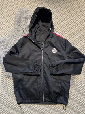 Svart Moncler vindjacka med huva - Svart vindjacka från Moncler med huva och dragkedja framtill. Jackan har röda, vita och blå detaljer på axlarna samt Moncler-logga på bröstet. Tillverkad i lätt och tåligt material, perfekt för blåsiga dagar. Justerbar nederkant med snören och två sidofickor.