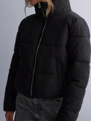 Svart pufferjacka med hög krage - Stilren svart pufferjacka med hög krage och dragkedja framtill. Jackan har en boxig passform och breda kanaler som ger en modern look. Perfekt för kalla dagar och enkel att matcha med olika outfits.