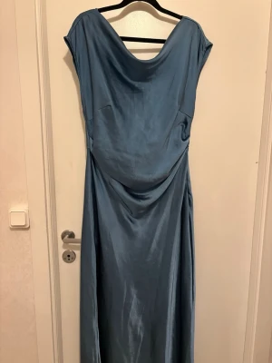 Blå turkos långklänning  - Snygg blå långklänning i glansigt material med draperad urringning bak. Klänningen har en elegant och flytande siluett, är ärmlös och passar perfekt för speciella tillfällen. Materialet ger en lyxig känsla och faller fint längs kroppen.