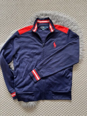 Marinblå cardigan tröja från Polo Ralph Lauren - Snygg marinblå cardigan med dragkedja från Polo Ralph Lauren. Har röda detaljer på axlar, krage och muddar samt det klassiska röda Polo-logot på bröstet. Tillverkad i mjukt bomullsmaterial och har sportig retrostil med ribbade muddar och krage.