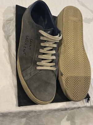 Grå Saint Laurent sneakers i mocka - Snygga grå sneakers från Saint Laurent i mjuk mocka med vit sula och vita snören. Diskret logga på sidan och klassisk låg modell som passar till det mesta. Insidan är fodrad i mörkblått tyg för extra komfort. Kom vid frågor, pris ej hugget i sten! Dessa är bara en tvätt ifrån att se nästan helt nya ut har dock inte haft energin att tvätta de.