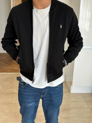 Svart bomberjacka från Ralph Lauren - Snygg svart bomberjacka från Ralph Lauren med klassisk passform och dragkedja framtill. Jackan har ribbade muddar vid ärmslut och midja samt den ikoniska loggan broderad på bröstet. Perfekt till jeans och t-shirt för en clean look.