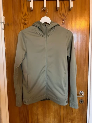 Grön hoodie från Peak Performance - Snygg ljusgrön hoodie från Peak Performance med dragkedja framtill och huva. Loggan syns på huvan och nedtill på tröjan. Mjuk och skön, perfekt för chill eller träning. Två fickor på sidorna och stilren design. Finns inga skavanker 