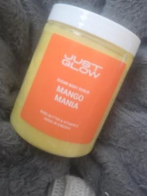Just Glow Mango Mania Body Scrub - Sockerbaserad kroppsskrubb från Just Glow med doft av mango. Innehåller sheasmör och vitamin E. 300g, oanvänd.  Katt finns i hemmet.  